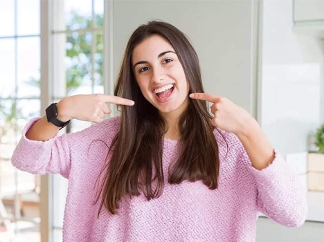 Woman in pink sweater pointing to her smile in bright, modern dental office setting.