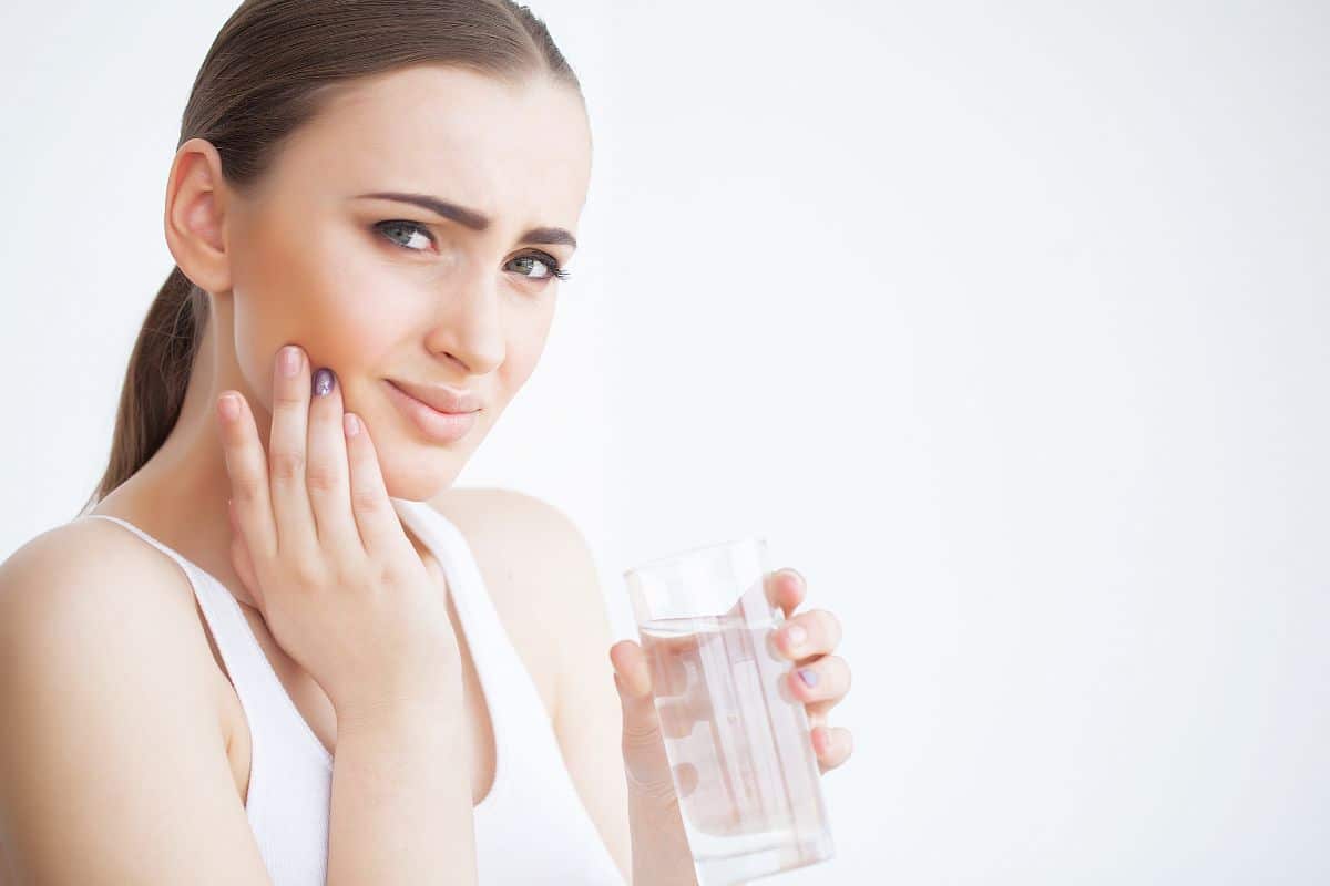 A young woman holds a glass of cold water and touches her cheek while experiencing tooth or gum pain.